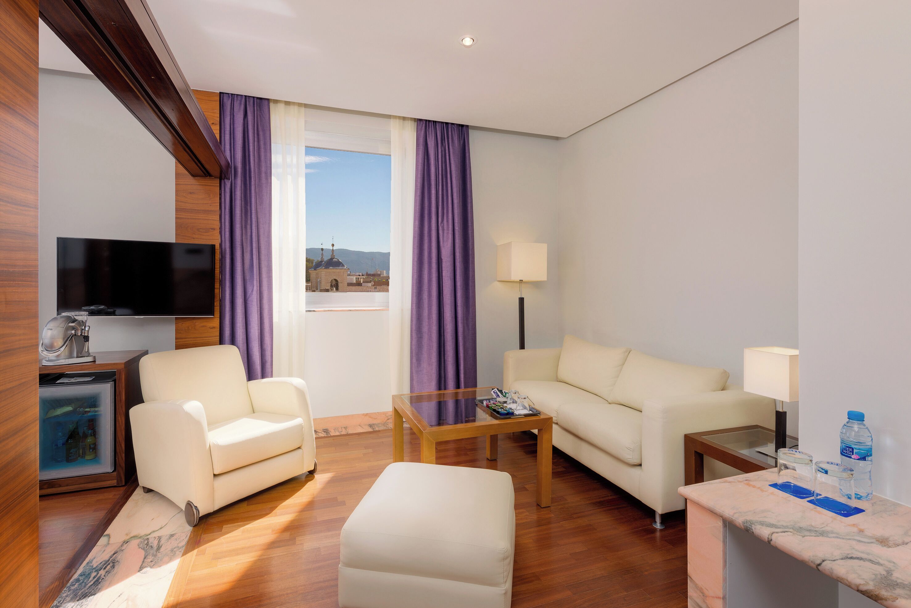 Ein Hotelzimmer mit Sofa, Sessel, Hocker, Couchtisch, Fernseher, Lampe, Minibar und Blick auf ein Gebäude durch das Fenster.
