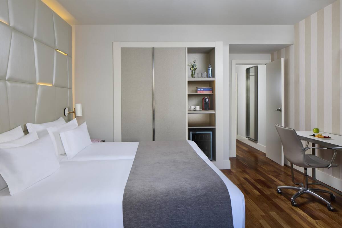 A hotel room with a bed, a desk, and a chair. There is a wardrobe and a shelf with items on it.