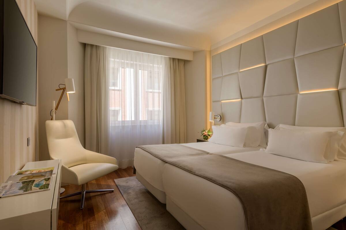 A modern hotel room with a large bed, a chair, a desk with a lamp, and a TV.