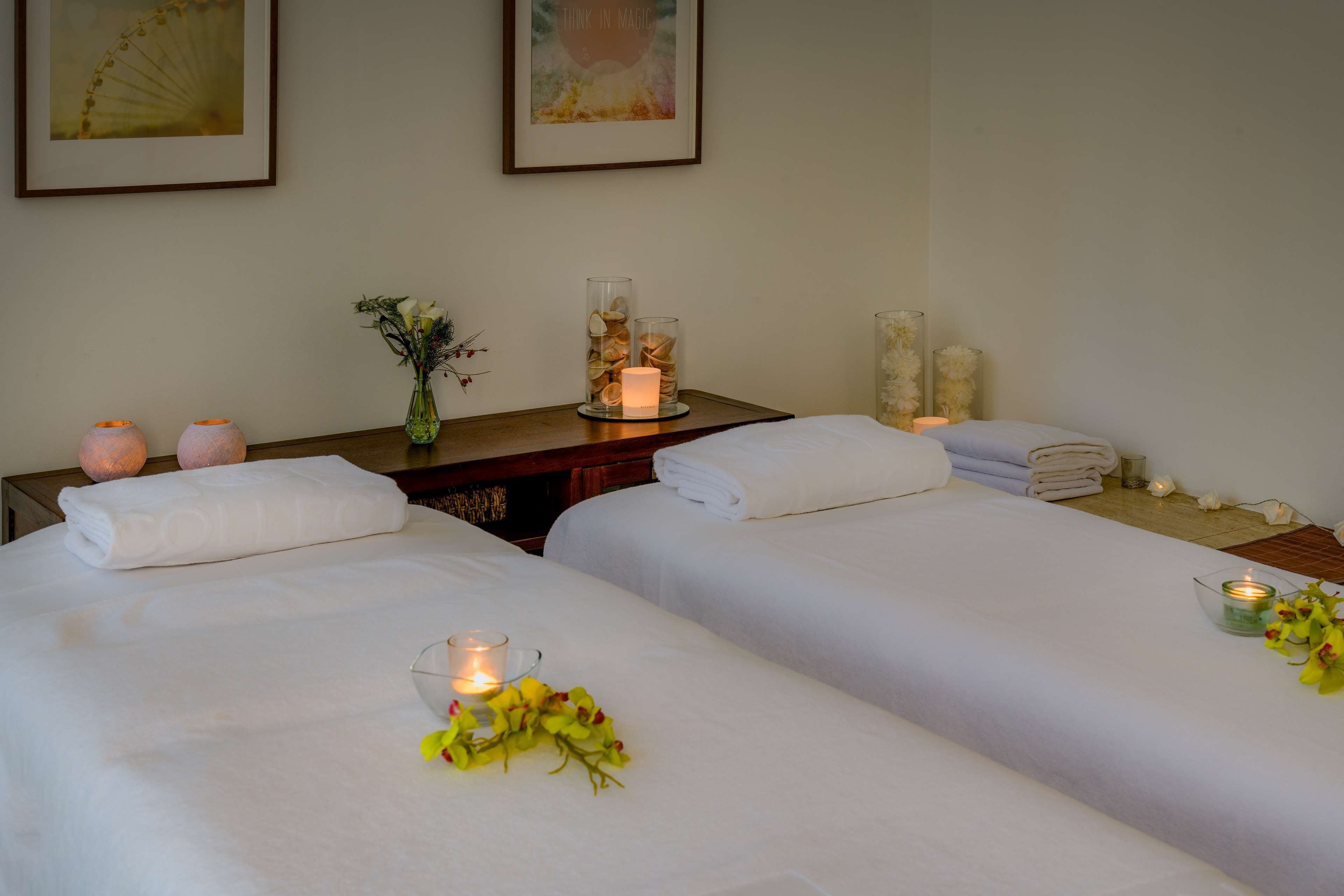 A hotel room with two beds, a bedside table with candles, a vase with flowers, and towels neatly stacked.
