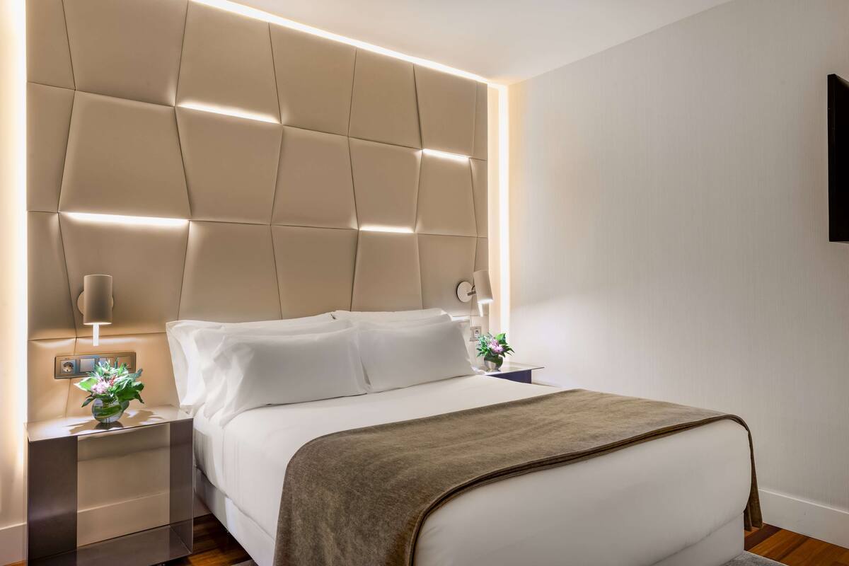 A modern hotel room with a tufted headboard, a bedside table with a lamp, and a decorative vase.