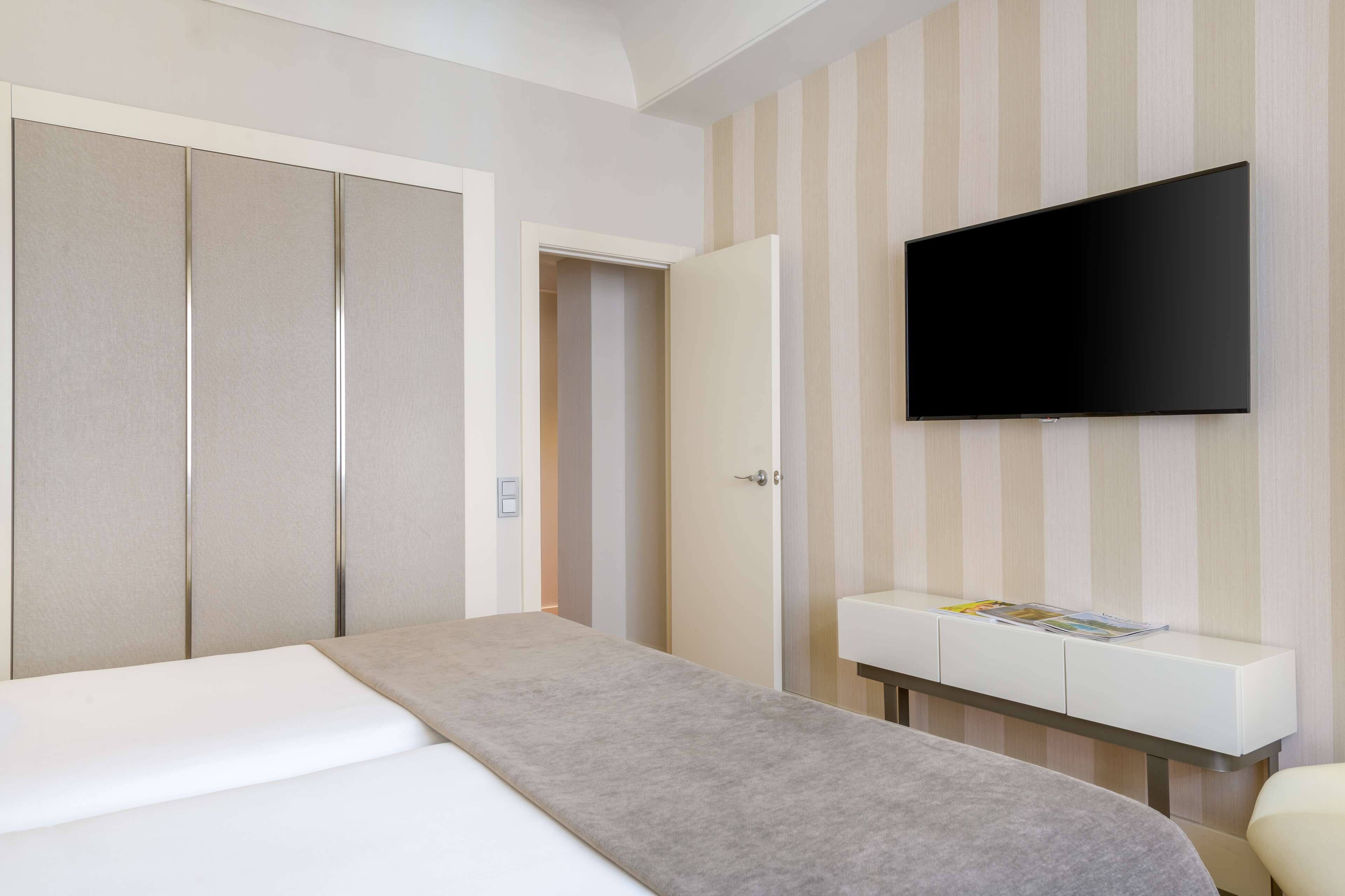 Habitación de hotel con cama, televisor montado en la pared, mesita de noche con una revista y armario.