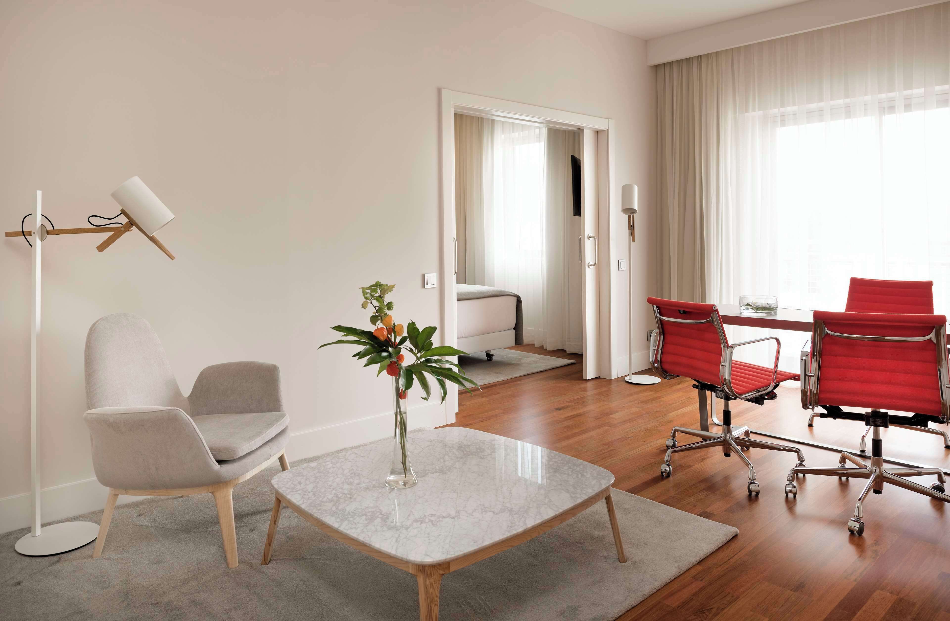 Quarto de hotel moderno com mesa de centro de mármore, poltrona cinza, cadeira de escritório vermelha e uma pequena planta.