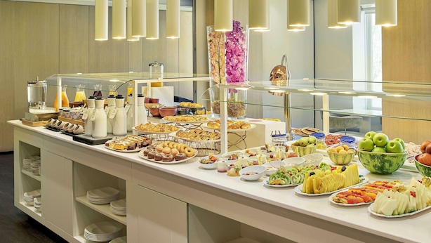 Un bufé con diversas opciones de comida y bebidas, incluyendo frutas, pastelería y una vitrina con flores.