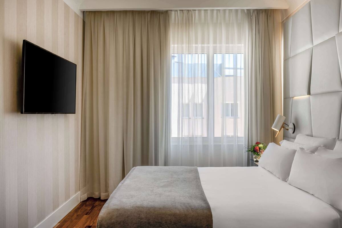 A hotel room with a large bed, a flat-screen TV mounted on the wall, sheer curtains, and a window with a view.