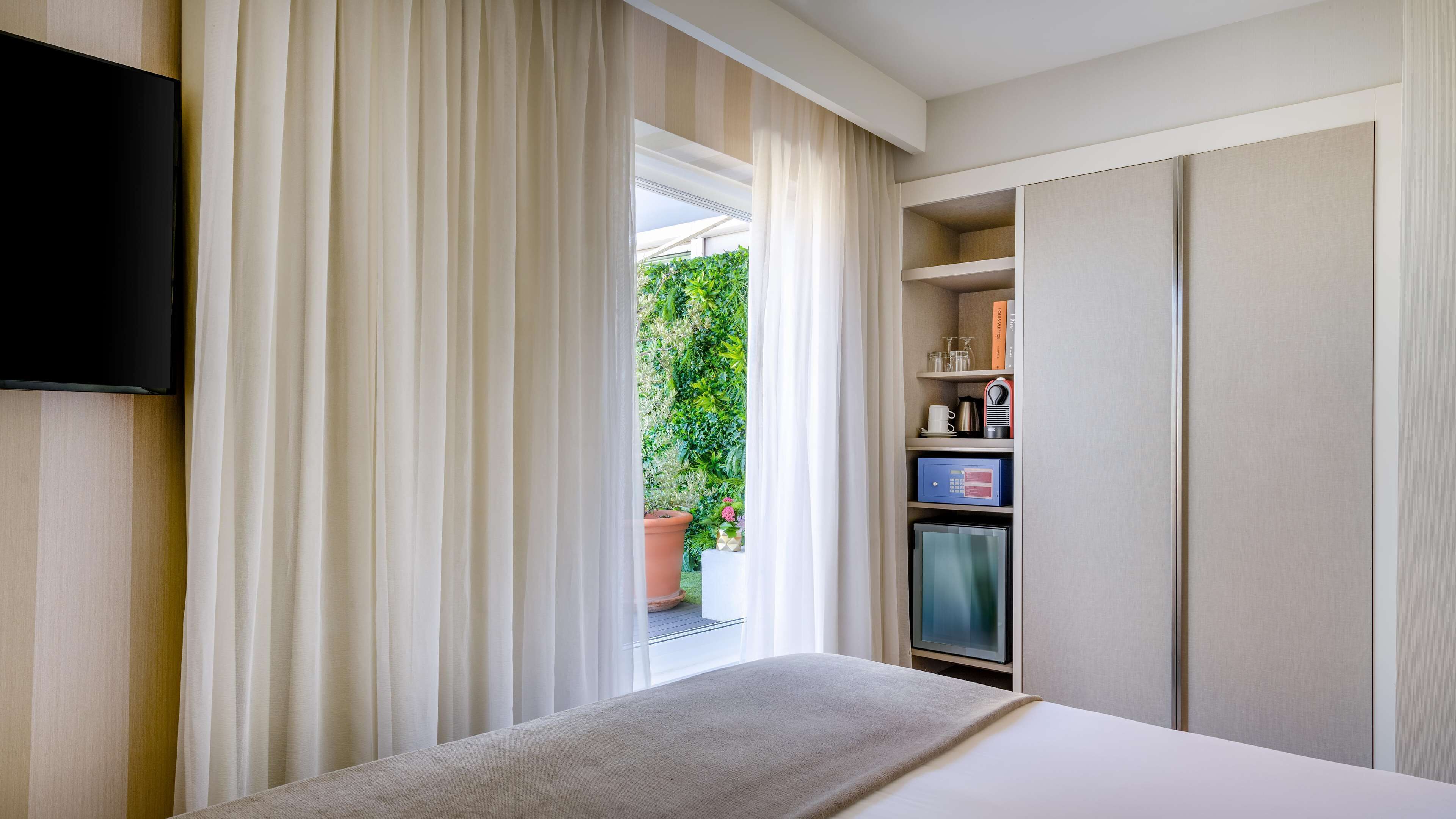 A hotel room with a bed, a TV mounted on the wall, a minibar, and a view of a garden through the window.