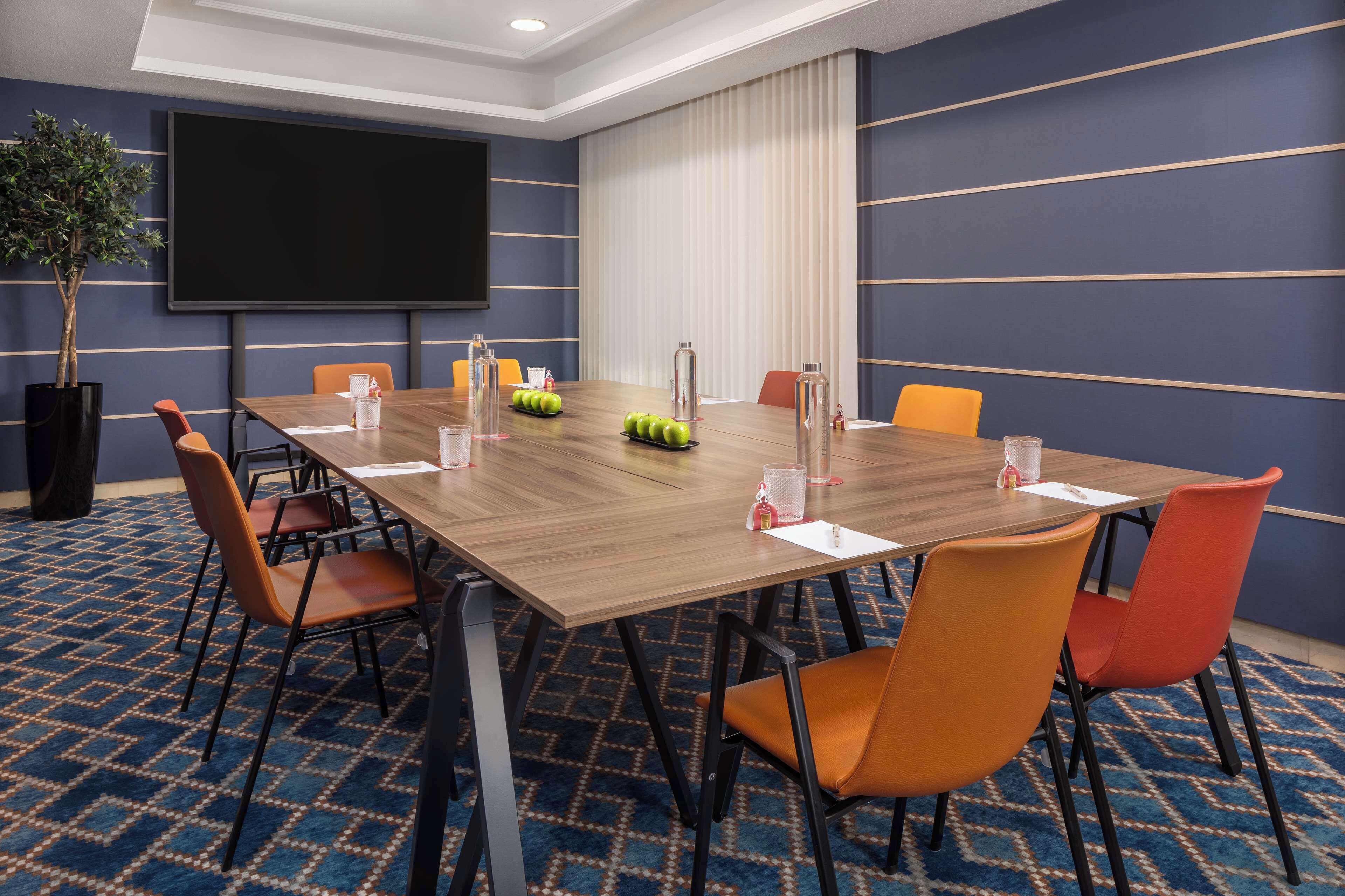 Ein Konferenzraum mit großem Tisch, orangefarbenen Stühlen, einer Projektionsleinwand und einem Blumentopf.