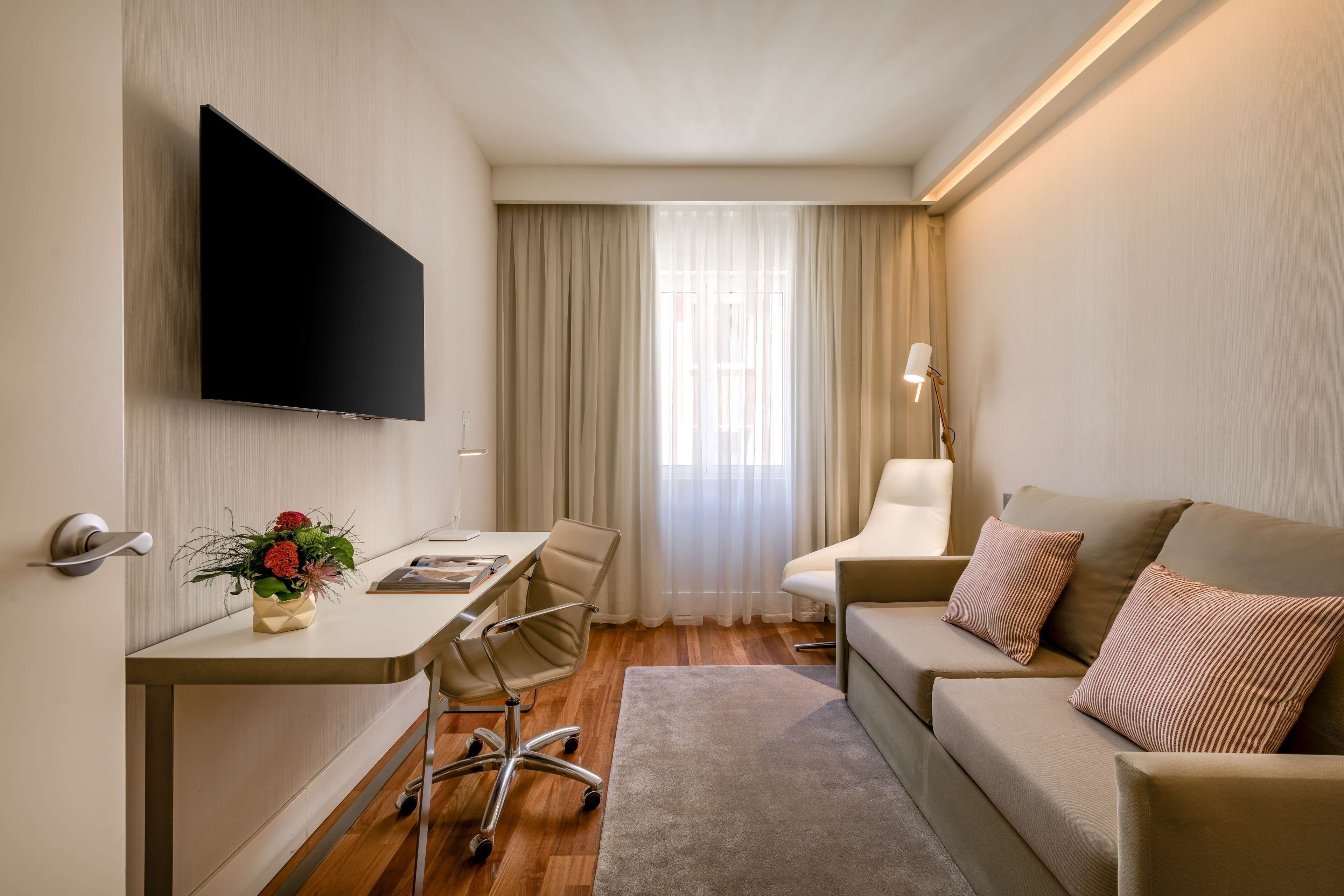 Una habitación de hotel moderna con televisor de pantalla plana, un escritorio con silla, un sofá y una lámpara.