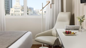 Habitación de hotel con cama, una silla, una mesa con un libro y una bandeja de frutas, y vista a una ciudad con una torre del reloj.