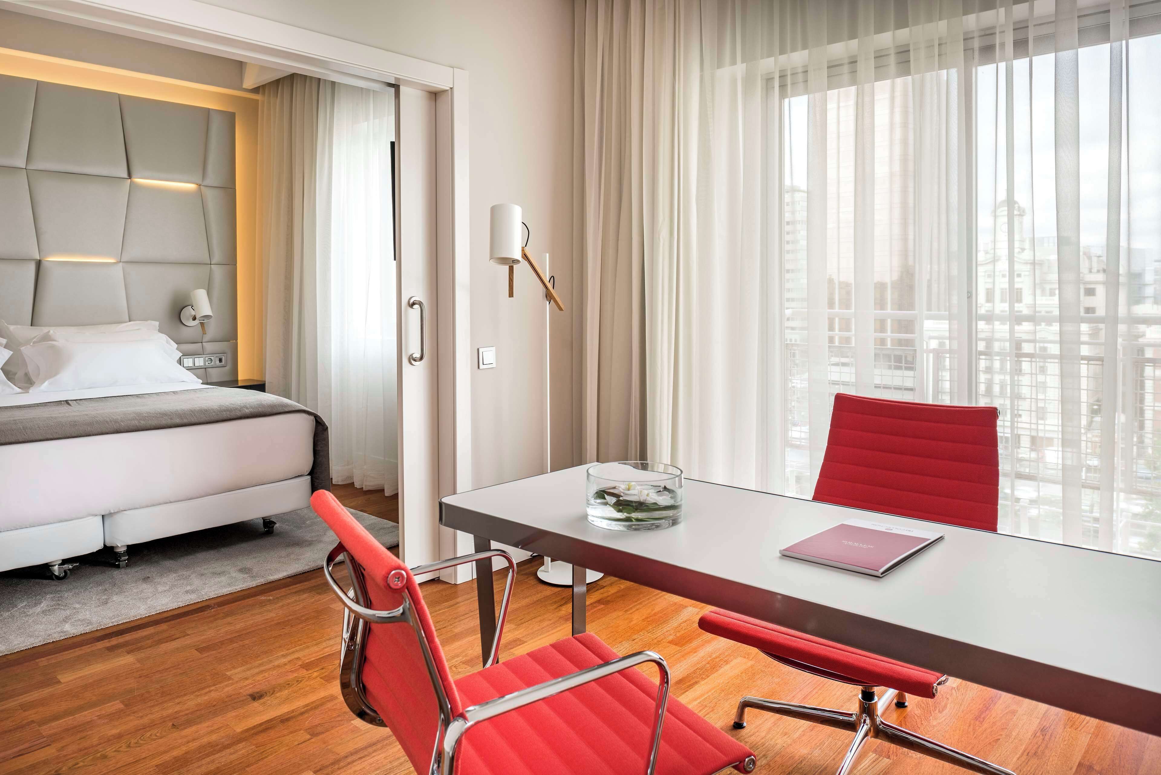 A hotel room with a bed, a glass table with a red chair, a city view, and a modern design.
