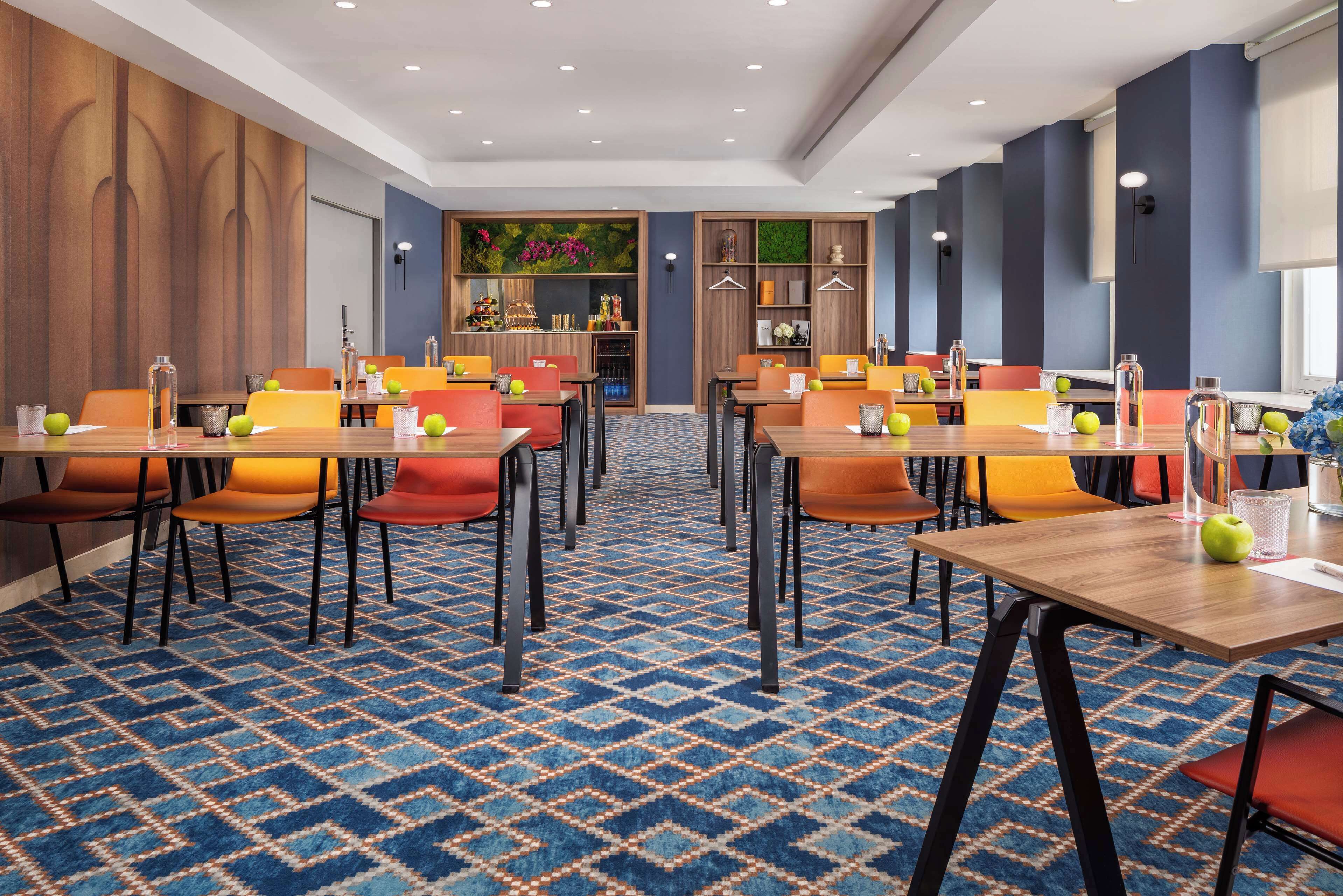 A hotel dining area with multiple tables and chairs, a patterned carpet, and a wall with a shelf displaying flowers and other items.