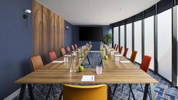 Un salón de conferencias con una larga mesa de madera, sillas anaranjadas y rojas, y una pantalla grande al frente.