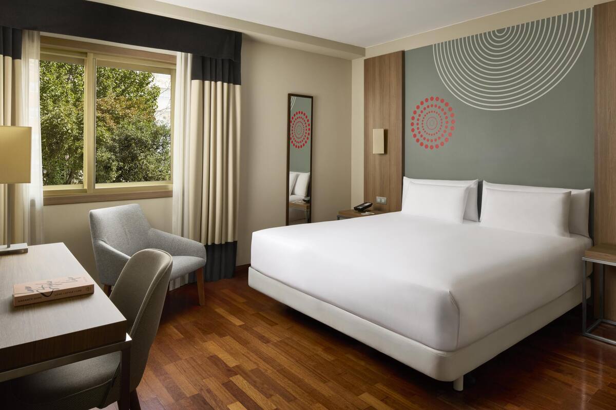 Une chambre d’hôtel avec un grand lit, un bureau avec une chaise, une fenêtre avec des rideaux et un mur orné de motifs circulaires.