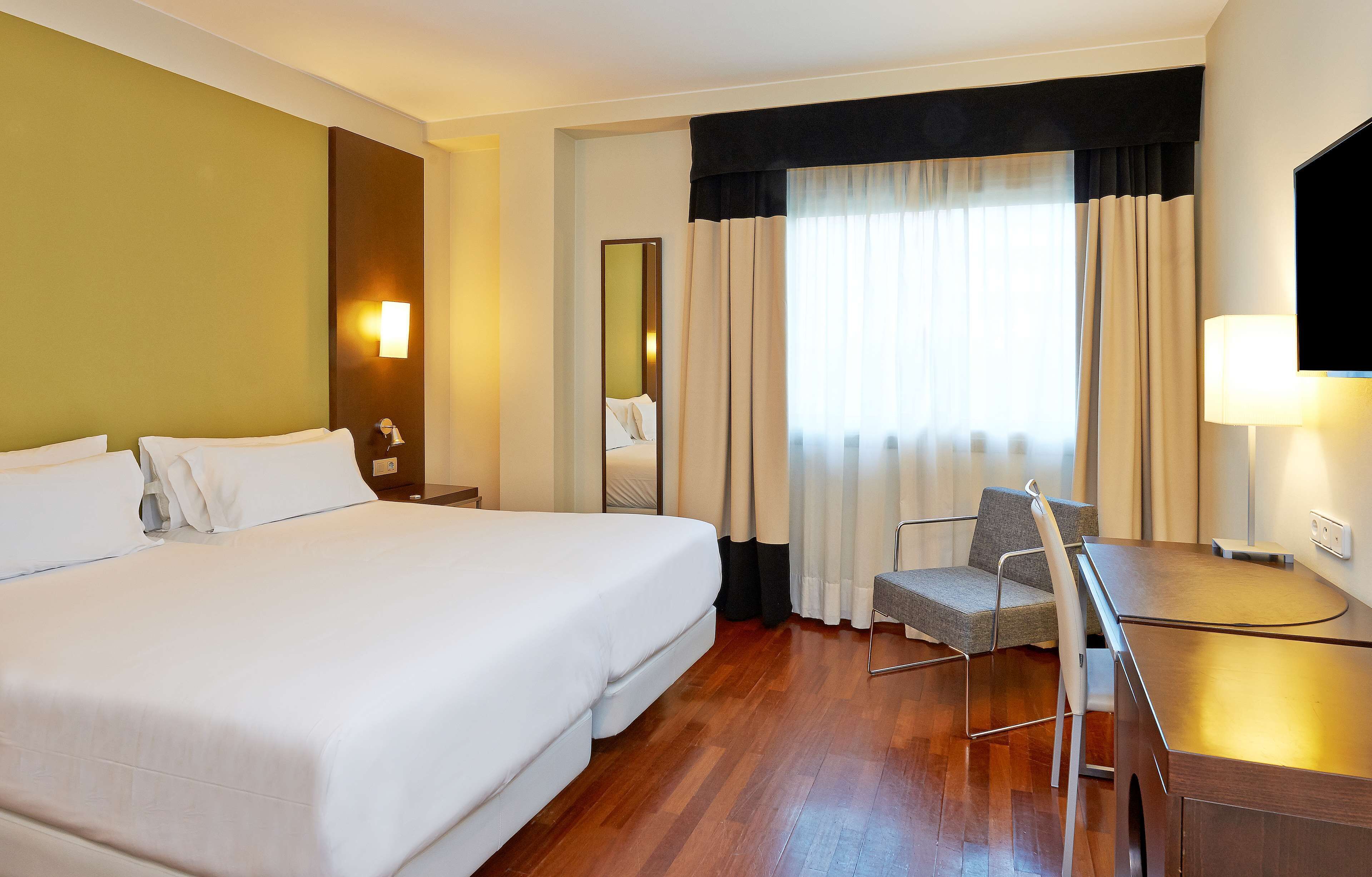 A hotel room with a large bed, a desk, a chair, a mirror, and a window with curtains.