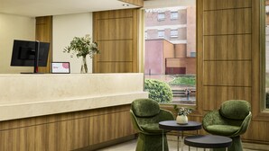 A reception area with a wooden reception desk, two green upholstered chairs, a small table, and a monitor.