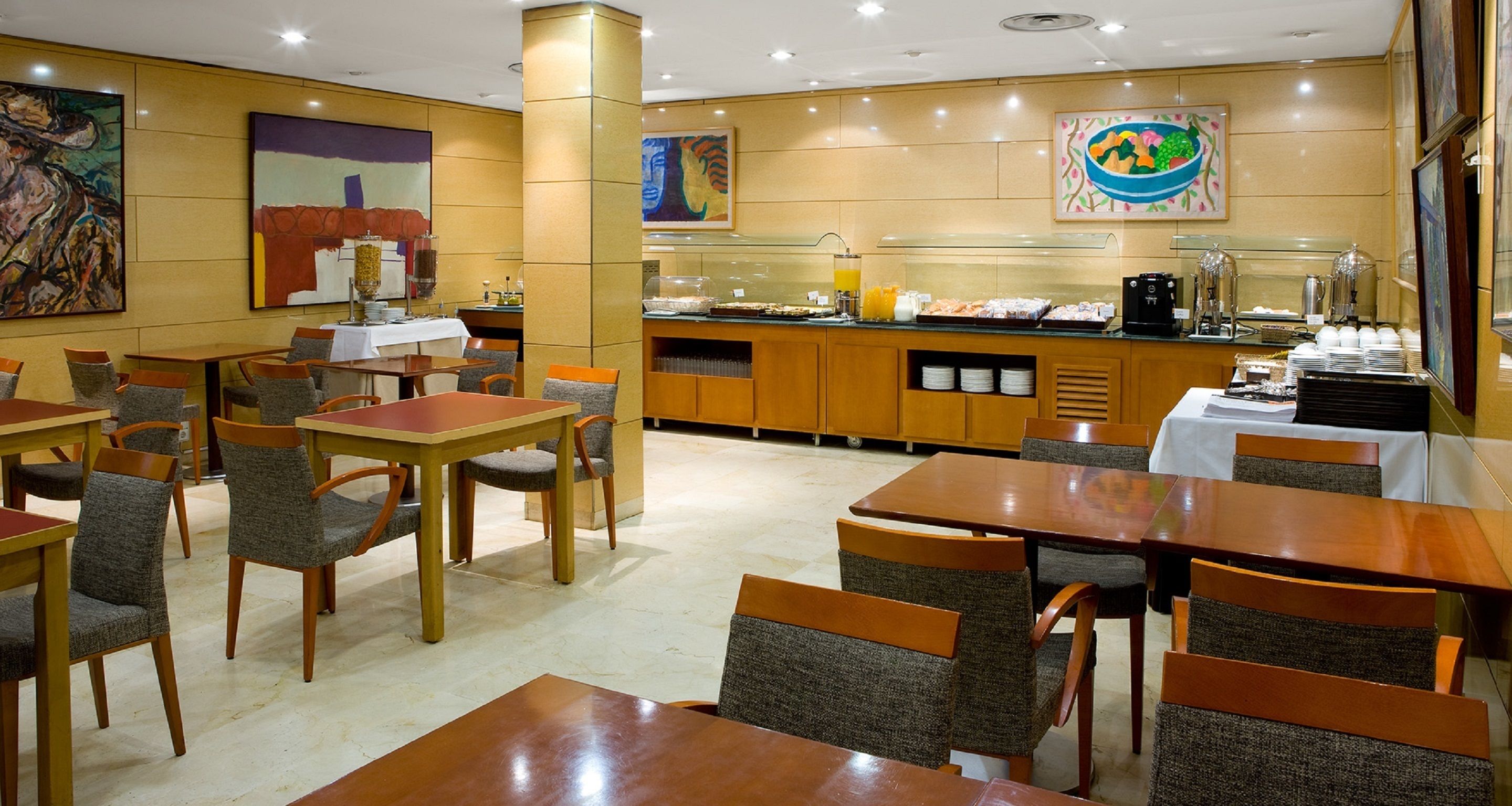 A hotel dining area with multiple tables and chairs, a buffet counter with food and beverages, and artwork on the walls.