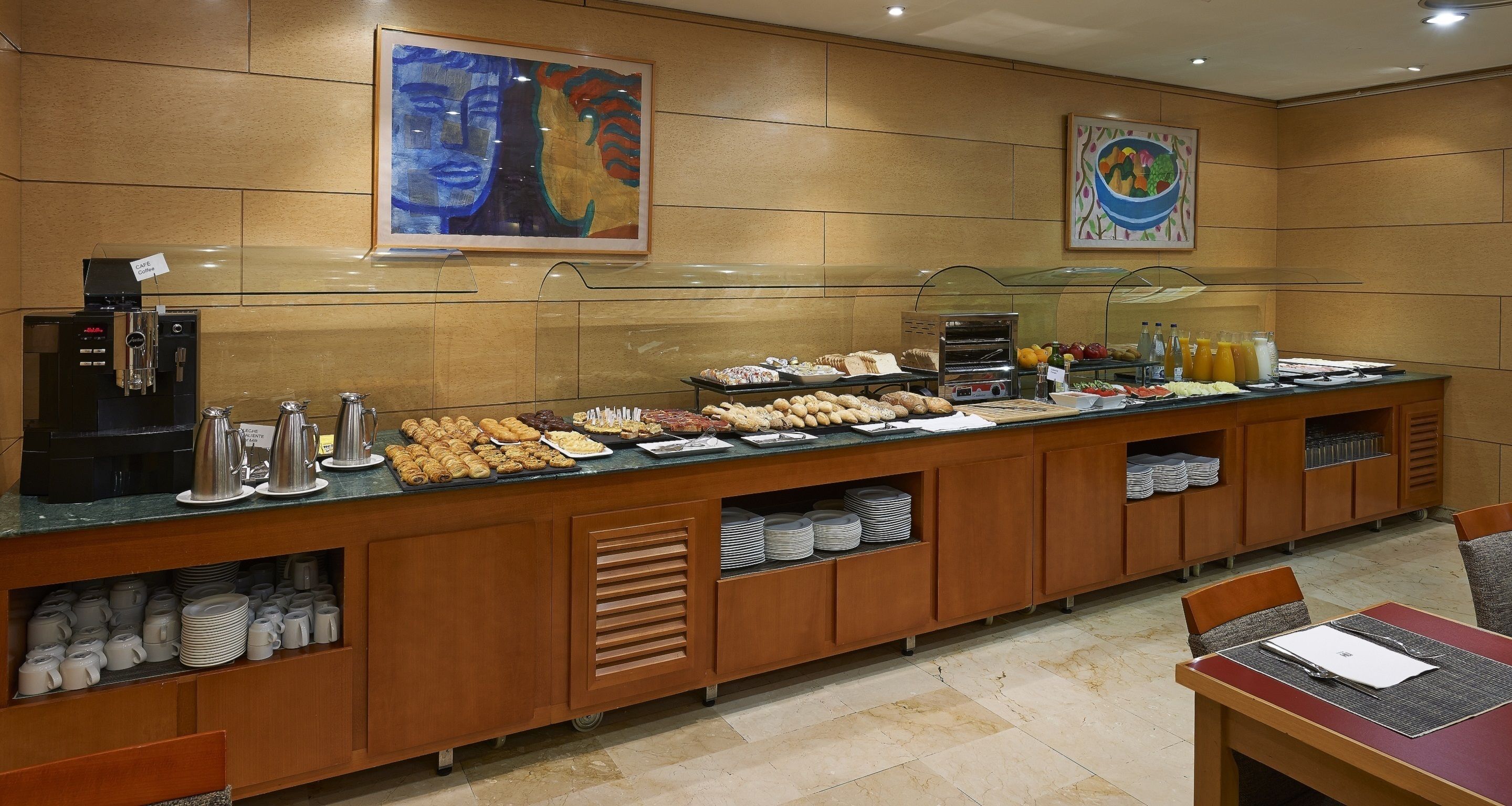 A buffet setup with various food items, a coffee machine, and a painting on the wall.