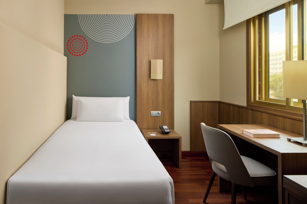 Une chambre d’hôtel avec un lit, un bureau, une chaise, une table de chevet, une lampe et une fenêtre.