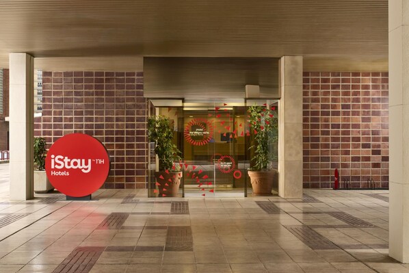 Entrada de hotel con logotipo rojo, puertas de vidrio, plantas en macetas y piso de baldosas.