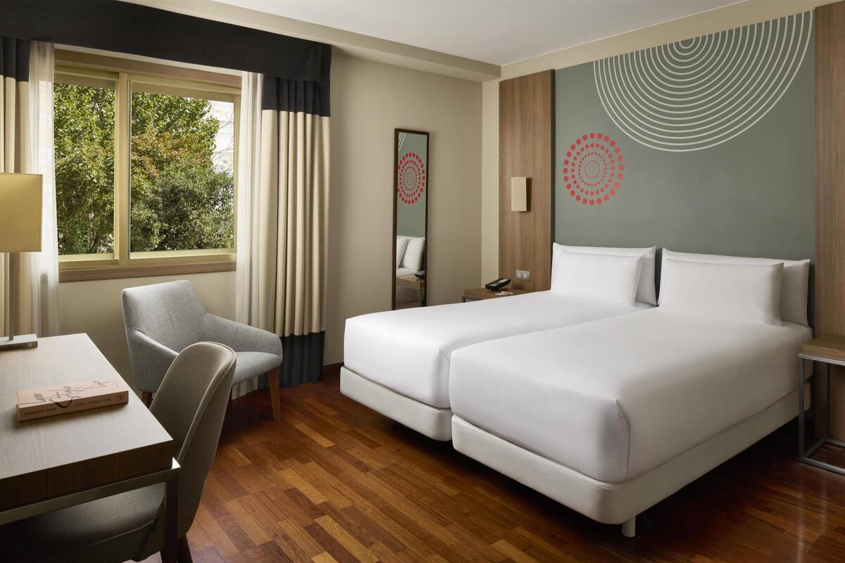 Une chambre d’hôtel avec un plancher en bois, un bureau avec une chaise, un lit avec deux oreillers, une table de chevet, un miroir et une fenêtre avec des rideaux.