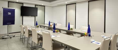 Una sala de conferencias con una mesa larga, sillas color beige, una pantalla de proyector y un televisor en la pared.