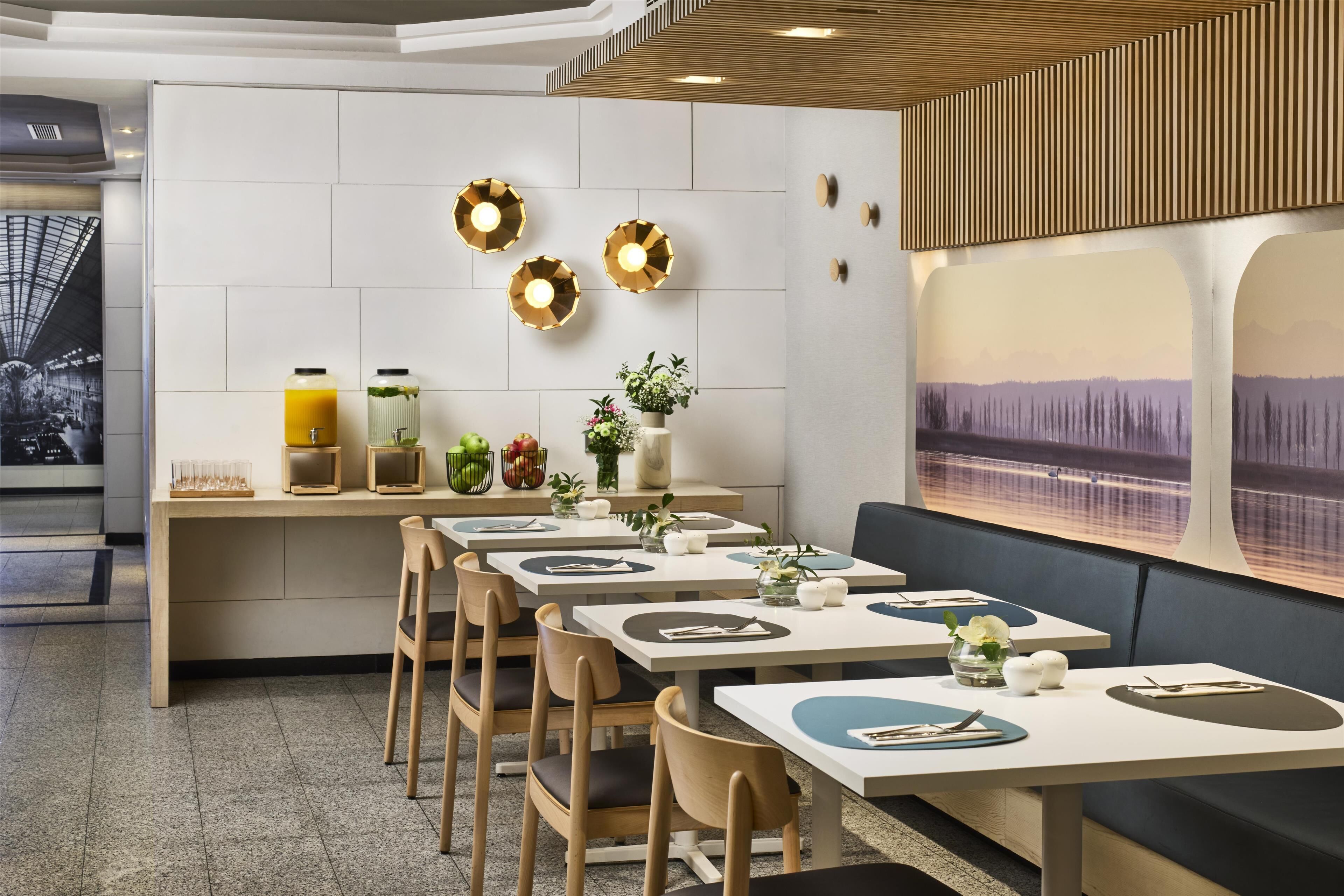 A modern dining area with wooden tables and chairs, a counter with food and drink containers, and decorative wall lights.