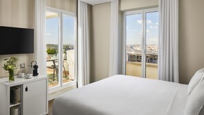 Ein Hotelzimmer mit einem großen Bett, einem an der Wand befestigten Fernseher, einem kleinen Schreibtisch mit Pflanze und Blick auf die Stadt durch große Fenster.