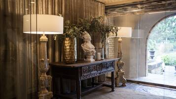 A stately room with a large arched window, a dark wooden console table with decorative lamps and vases, and a stone bust sculpture.