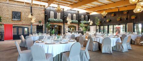 Un salón de banquetes con mesas redondas dispuestas para comer, una pared de piedra y vigas de madera en el techo.
