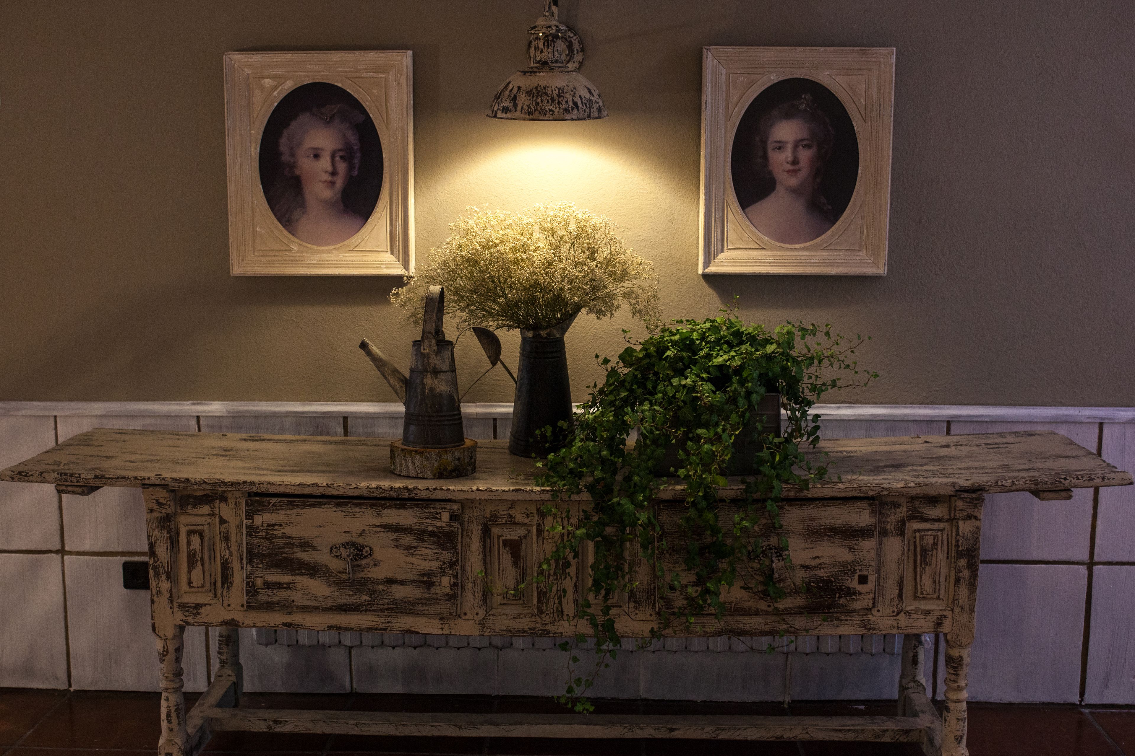 A rustic wooden table with a vase of flowers and a potted plant, set against a wall with framed portraits.
