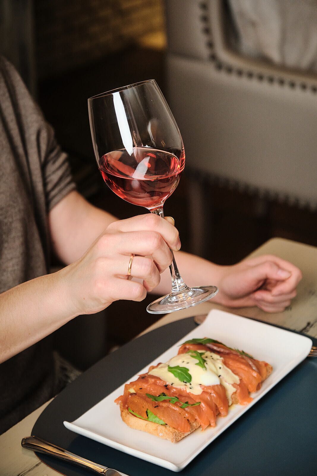 Una persona sosteniendo una copa de vino con líquido rojo, sentada a una mesa con un plato de comida.