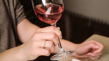 Una persona sosteniendo una copa de vino con líquido rojo, sentada a una mesa con un plato de comida.