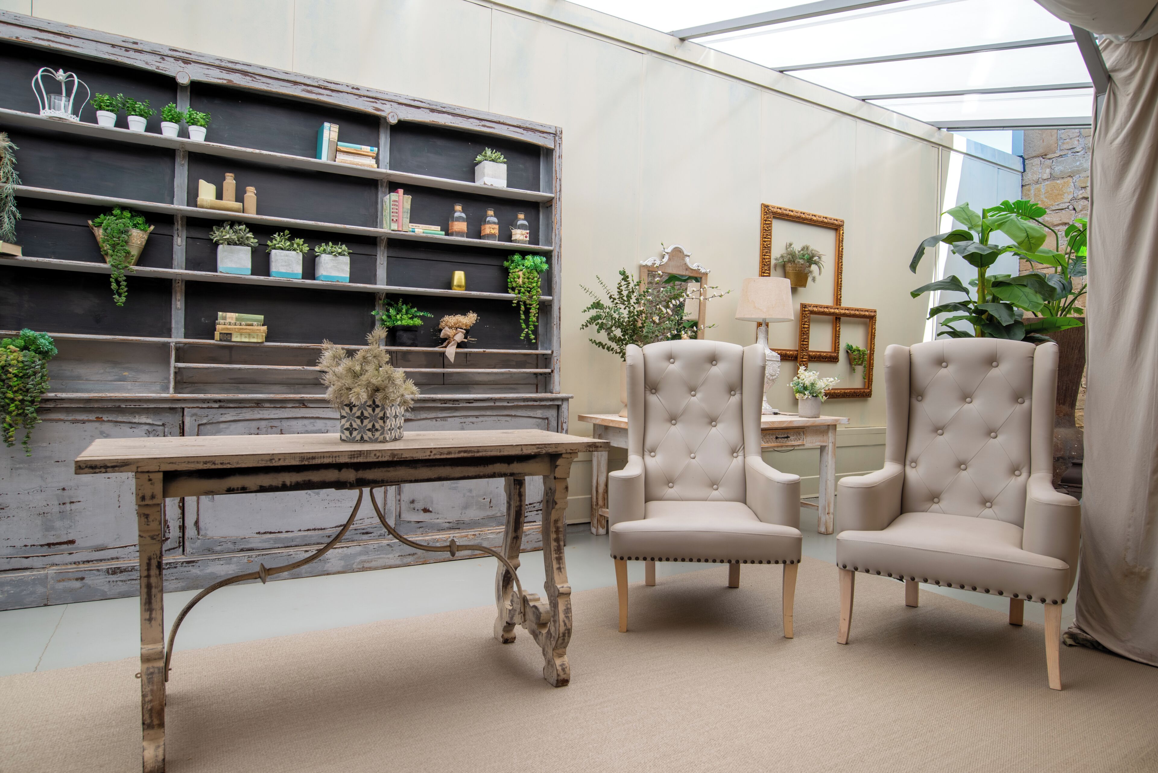 Una habitación con una mesa de madera rústica, dos sillones tapizados, una estantería con plantas y un espejo.