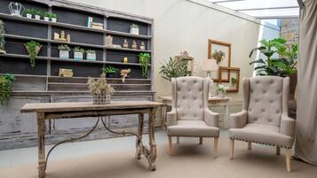 Una habitación con una mesa de madera rústica, dos sillones tapizados, una estantería con plantas y un espejo.
