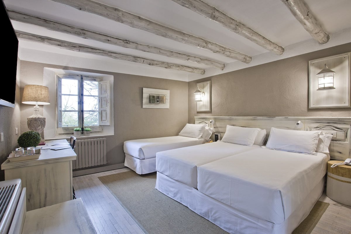 A bedroom with two beds, a window with shutters, a radiator, a lamp, and a small plant.