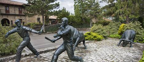 A statue of a man with a baseball bat and a cow in a courtyard with cobblestone flooring and surrounding greenery.