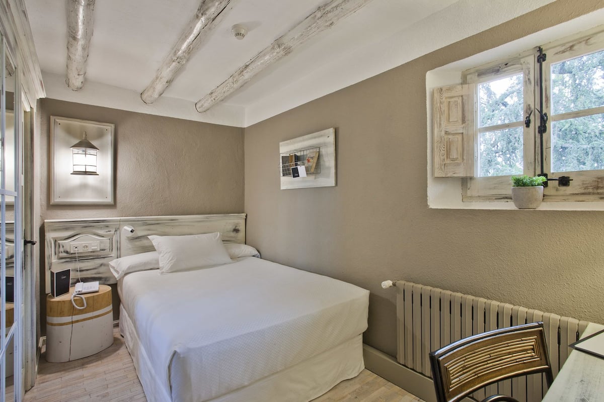 A bedroom with a bed, bedside table, lamp, radiator, window with curtains, and a small table with a chair.