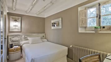 Un dormitorio con cama, mesita de noche, lámpara, radiador, ventana con cortinas y una pequeña mesa con una silla.