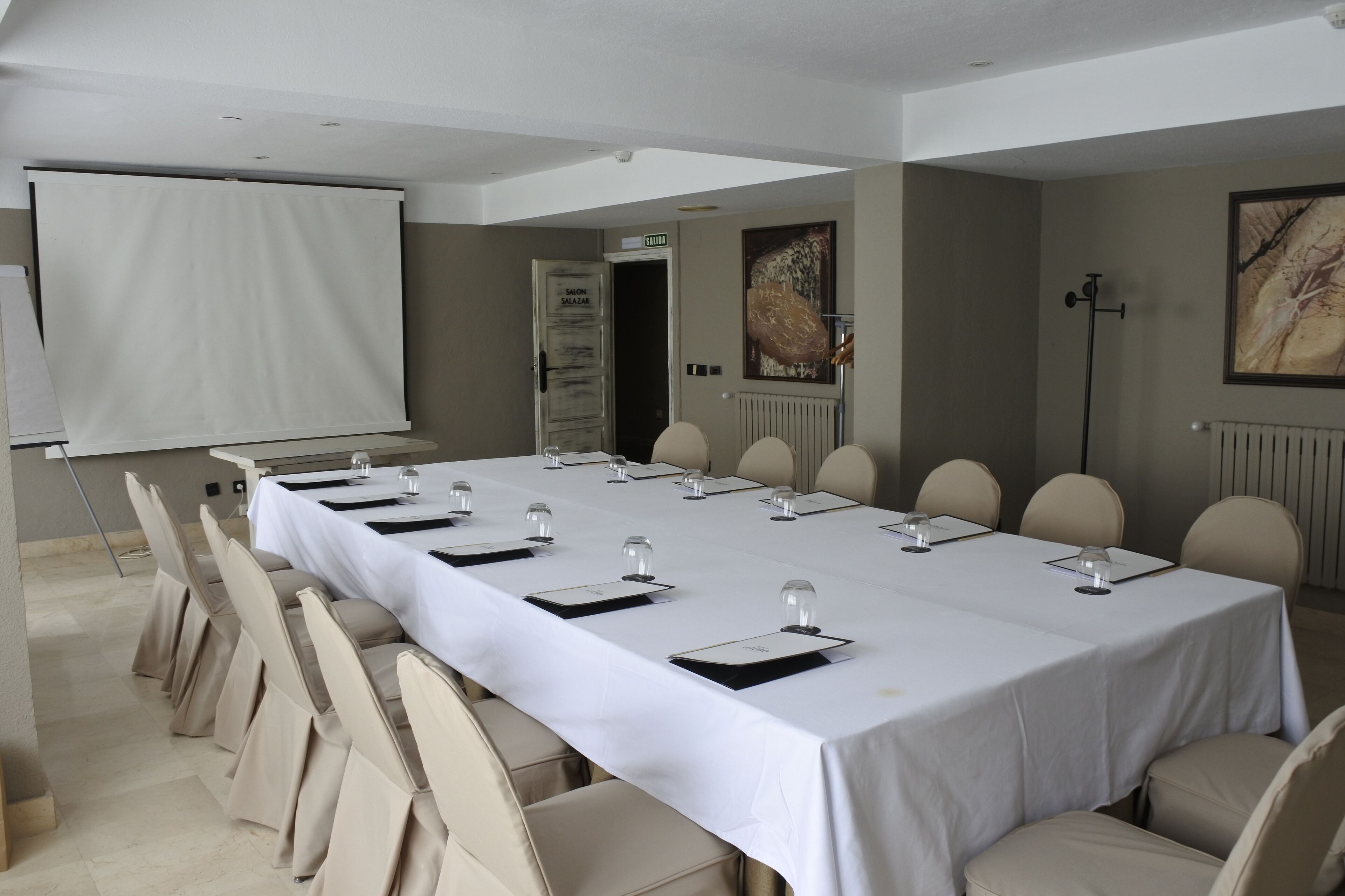 A conference room with a long table, chairs, a projector screen, and a door leading to a salon.