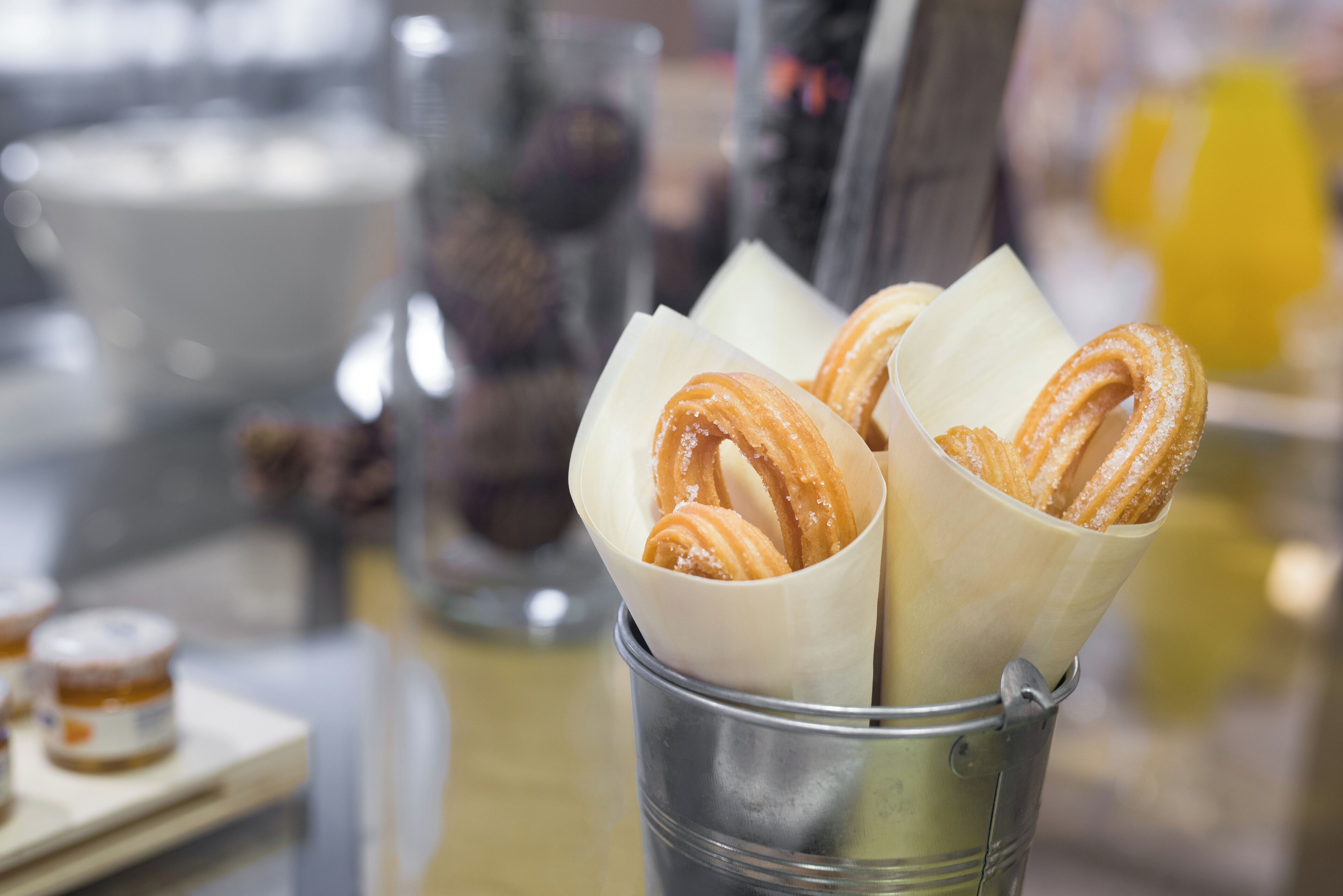 A metal cup filled with churros, a glass of milk, and a jar of jam on a table.