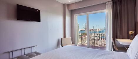 Habitación de hotel con una cama grande, un televisor de pantalla plana montado en la pared, un balcón con vistas a un puerto deportivo y una pequeña mesa con un florero.