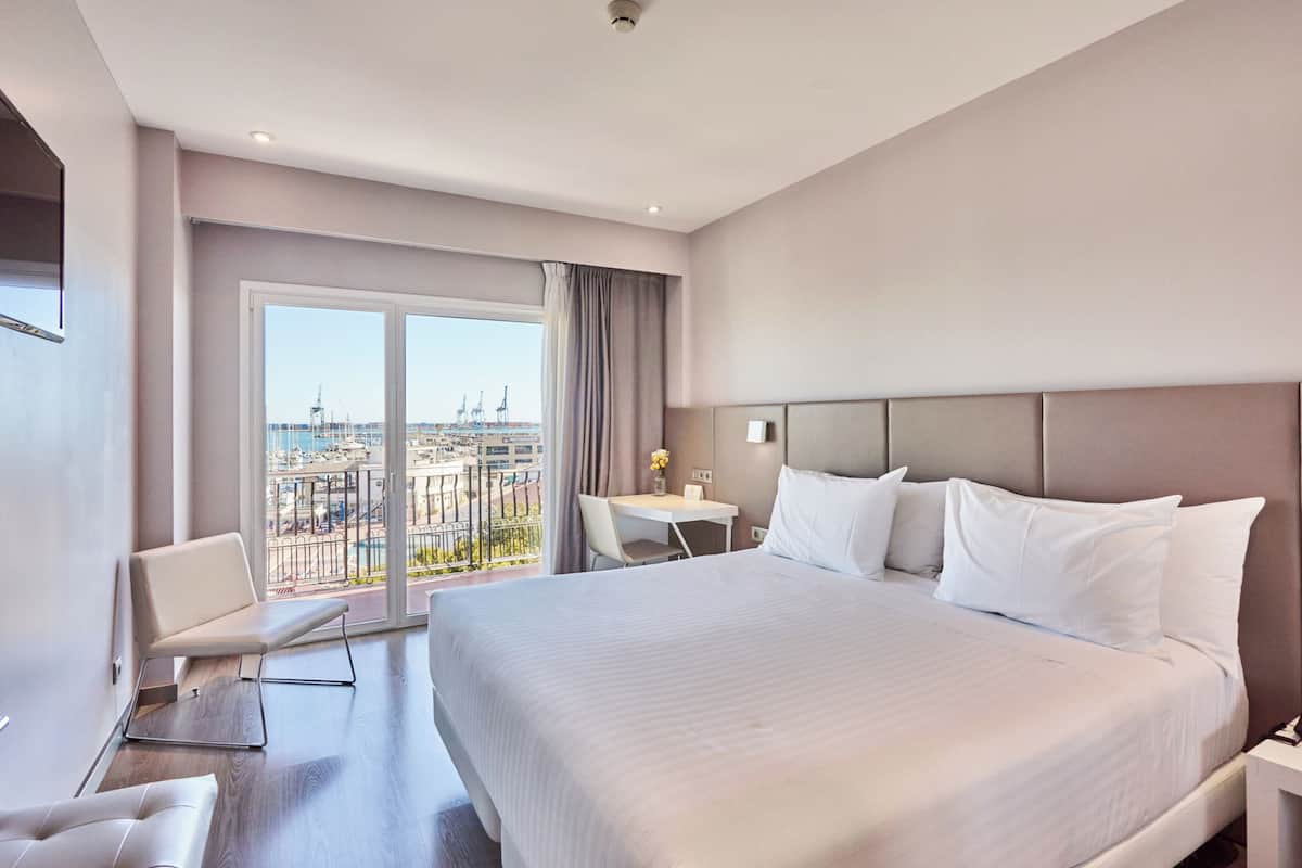 A hotel room with a large bed, a desk, and a chair. There is a view of the city through the window.