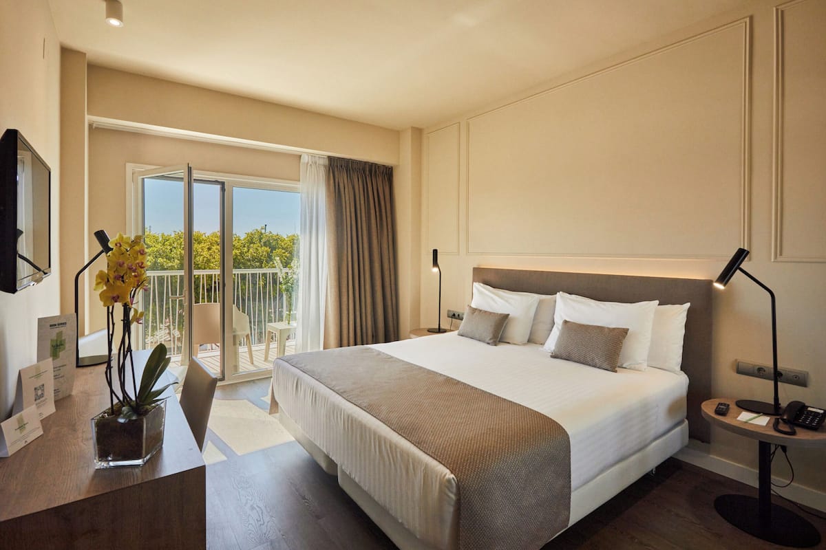 A hotel room with a large bed, a desk with a lamp, a small table with a phone, and a view of the outdoors.