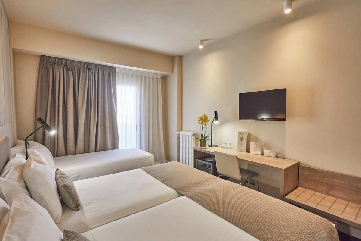 A hotel room with two beds, a desk, a TV, and a window with curtains.