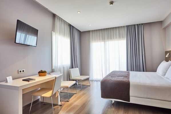 A modern hotel room with a bed, desk, and chair, large windows with curtains, and a flat-screen TV mounted on the wall.