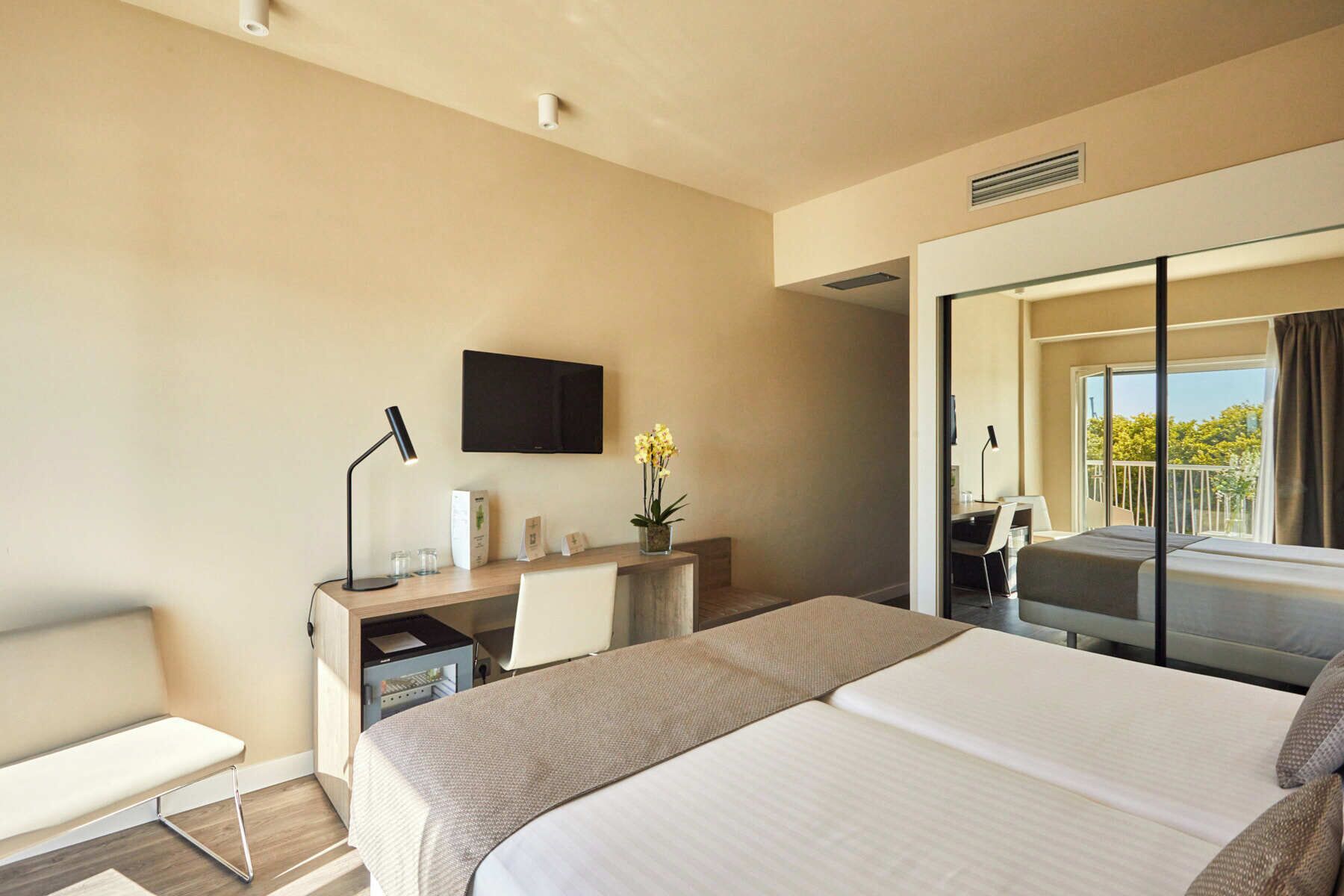 A hotel room with a bed, a desk with a chair, a TV mounted on the wall, and a balcony with a view.