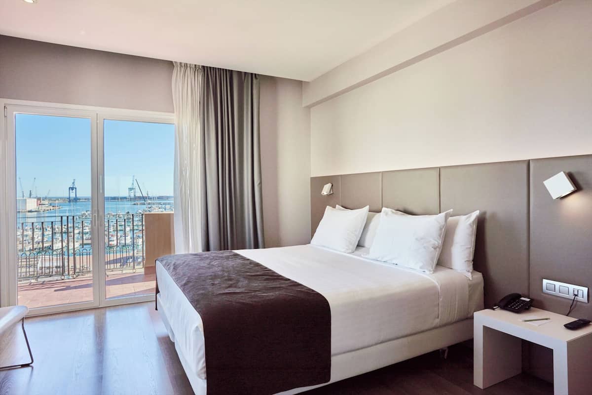 A hotel room with a large bed, a bedside table with a phone, and a view of the sea and port through a sliding door.