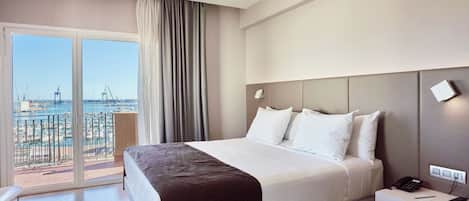 A hotel room with a large bed, a bedside table with a phone, and a view of the sea and port through a sliding door.