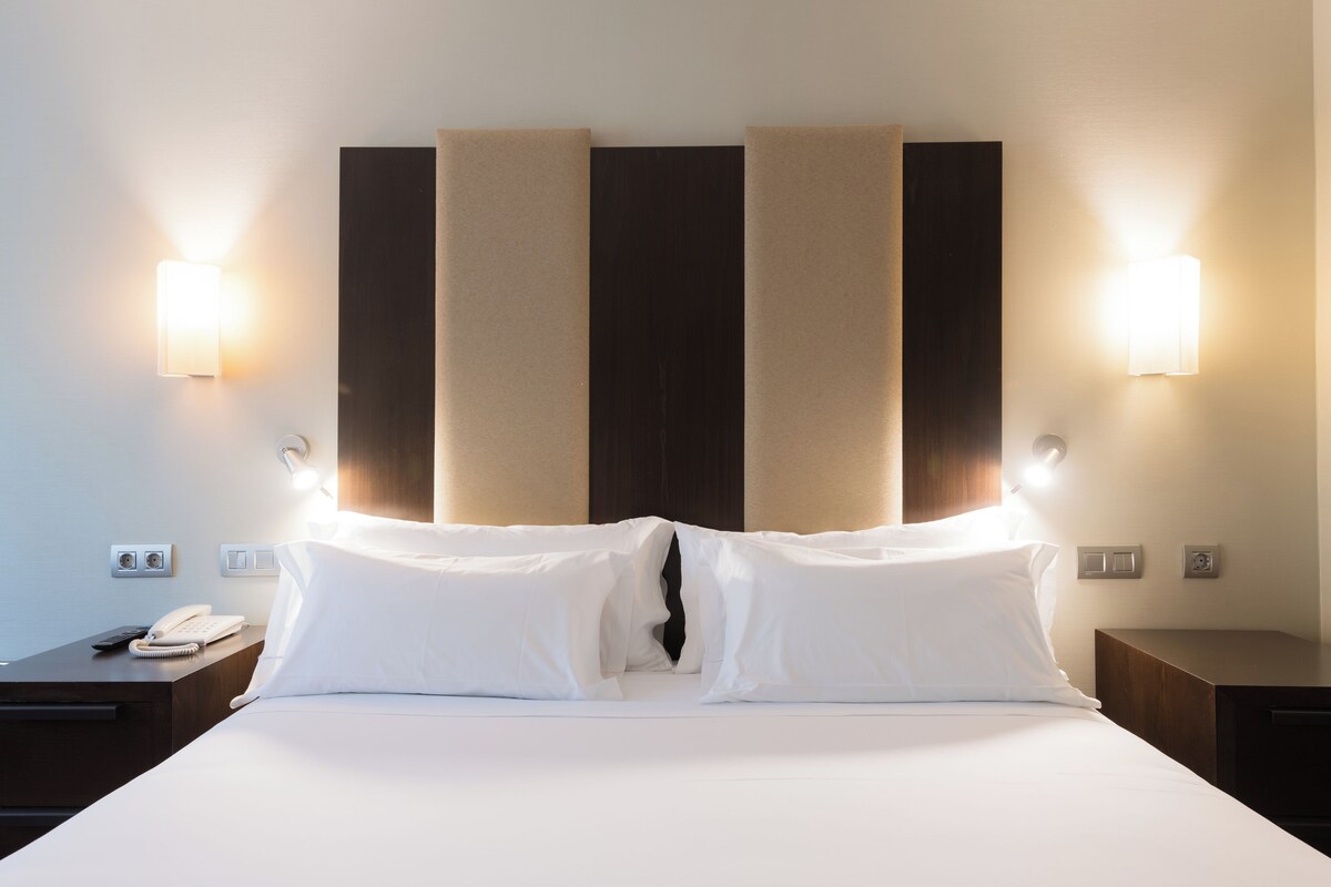 A hotel room with a large bed, two bedside tables, a nightstand, a phone, and wall-mounted lights.