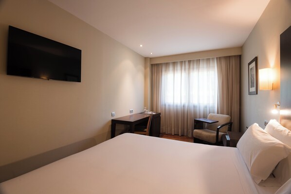 Habitación de hotel con una cama grande, un escritorio, una silla, un televisor de pantalla plana montado en la pared y una ventana con cortinas.